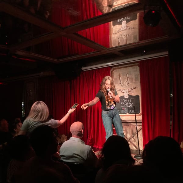 A female comedian performs on stage at The White Bull Tavern, with a crowd watching.