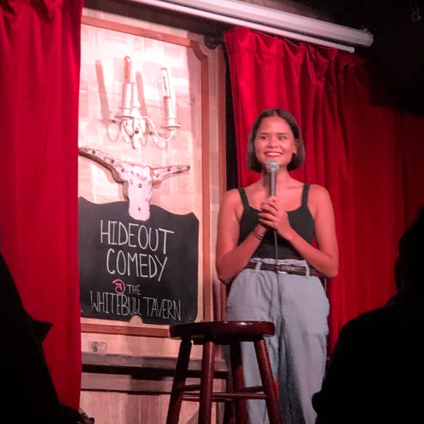 A woman performing stand-up comedy at The White Bull Tavern, with a "Hideout Comedy" sign.