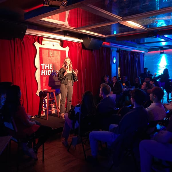A female comedian performs on stage with a microphone for an audience at The White Bull Tavern.