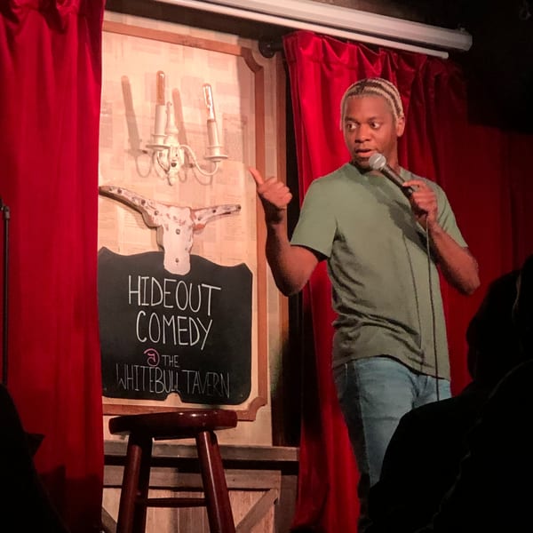 A comedian performs on stage at The White Bull Tavern, with a "Hideout Comedy" sign behind him.
