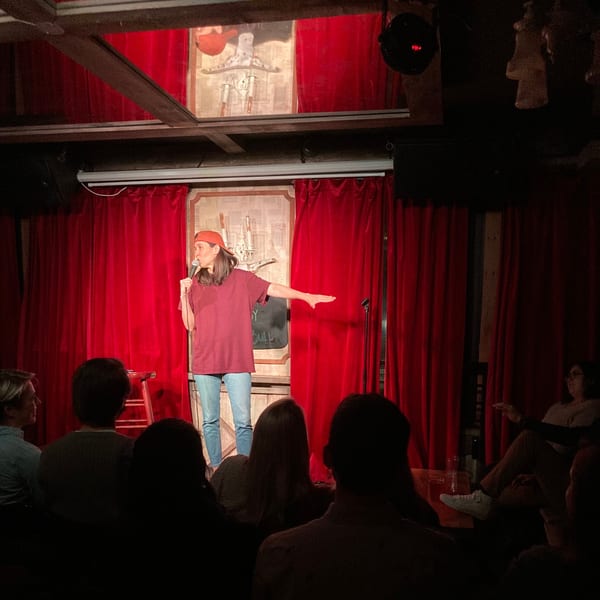 A comedian in a red shirt and cap performs on stage with red curtains at The White Bull Tavern.