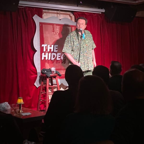 A comedian performs on stage at The White Bull Tavern, with red curtains and a sign.
