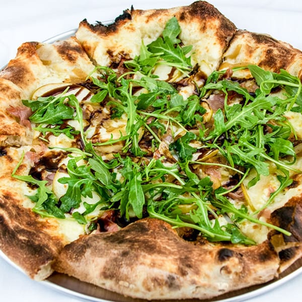 A close-up of a pizza topped with arugula, prosciutto, pears, and balsamic glaze.