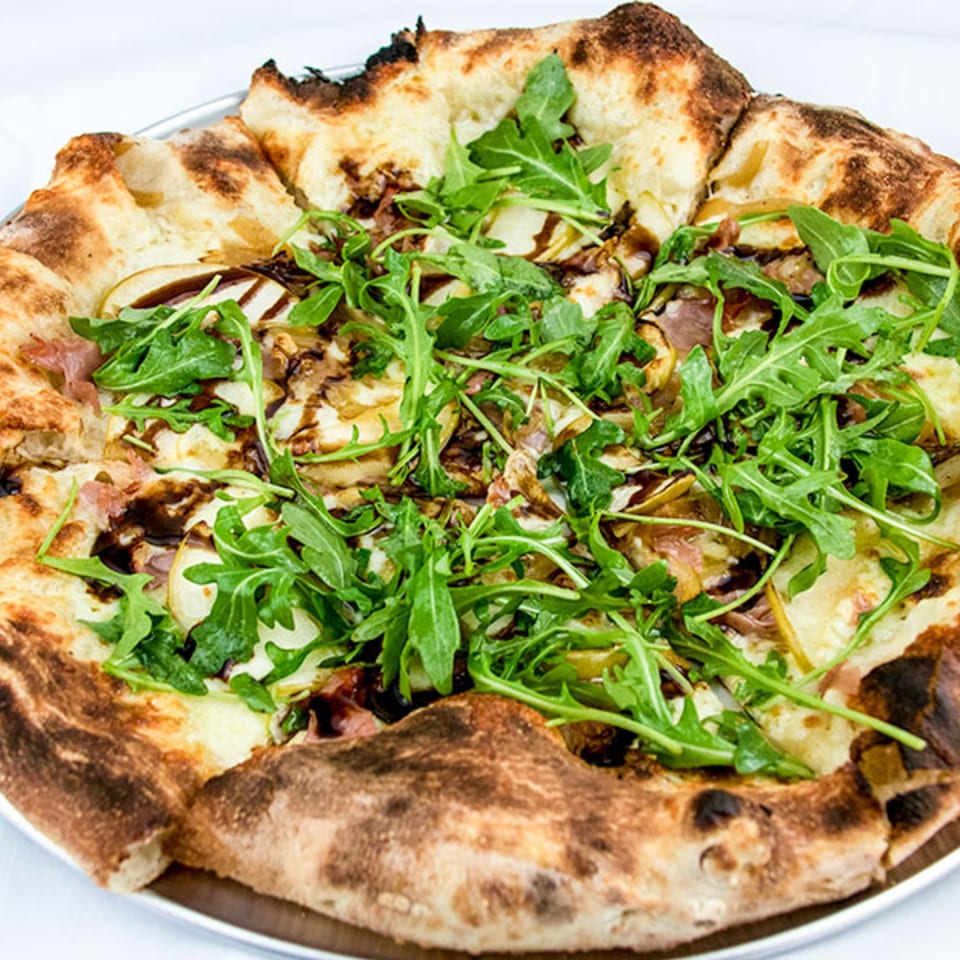 A close-up of a pizza topped with arugula, prosciutto, pears, and balsamic glaze.